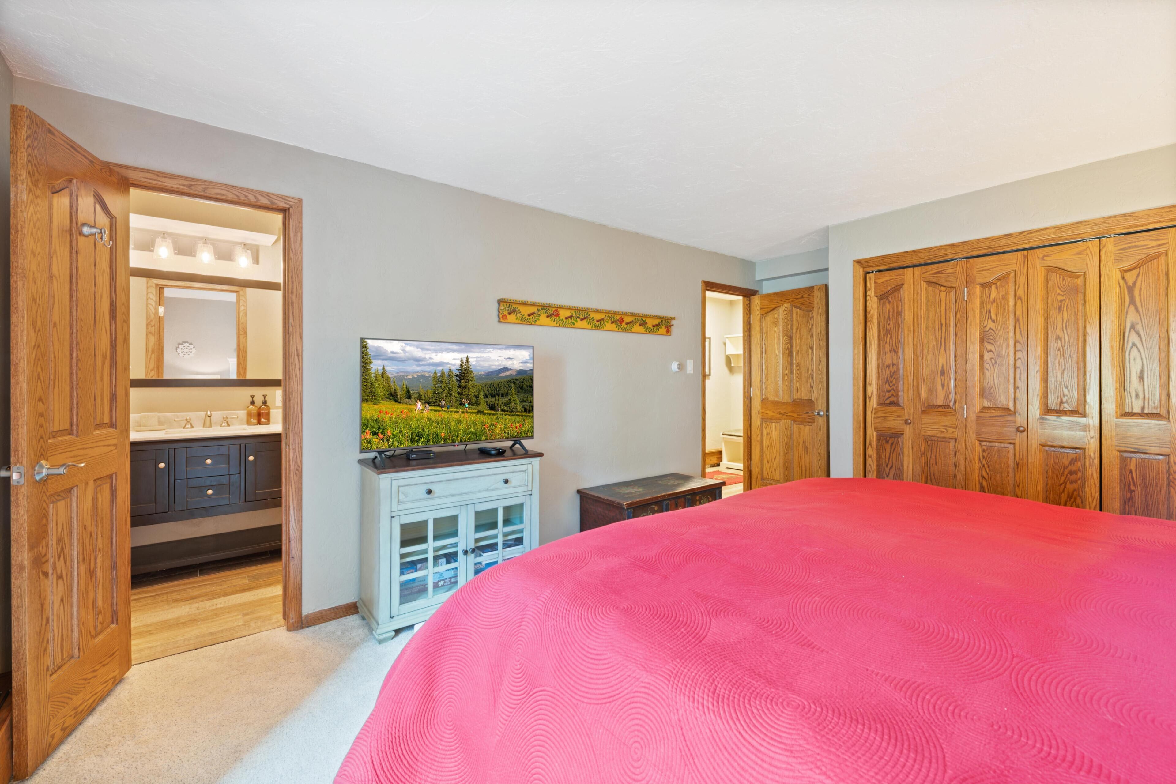 A bedroom with a large bed, wooden wardrobe, a TV, and a bathroom with a mirror and sink.