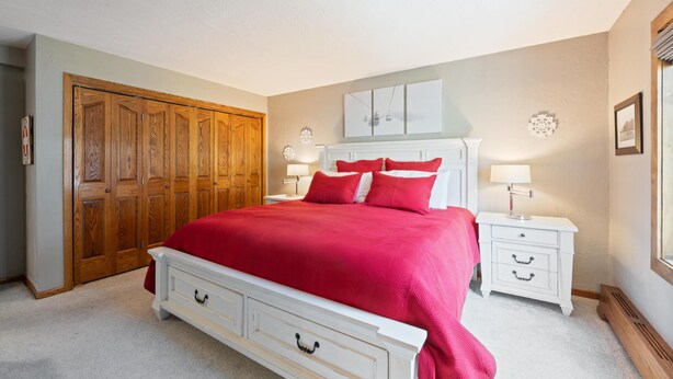 Quarto com cama grande, guarda-roupa de madeira, mesa de cabeceira com abajur e obra de arte na parede.