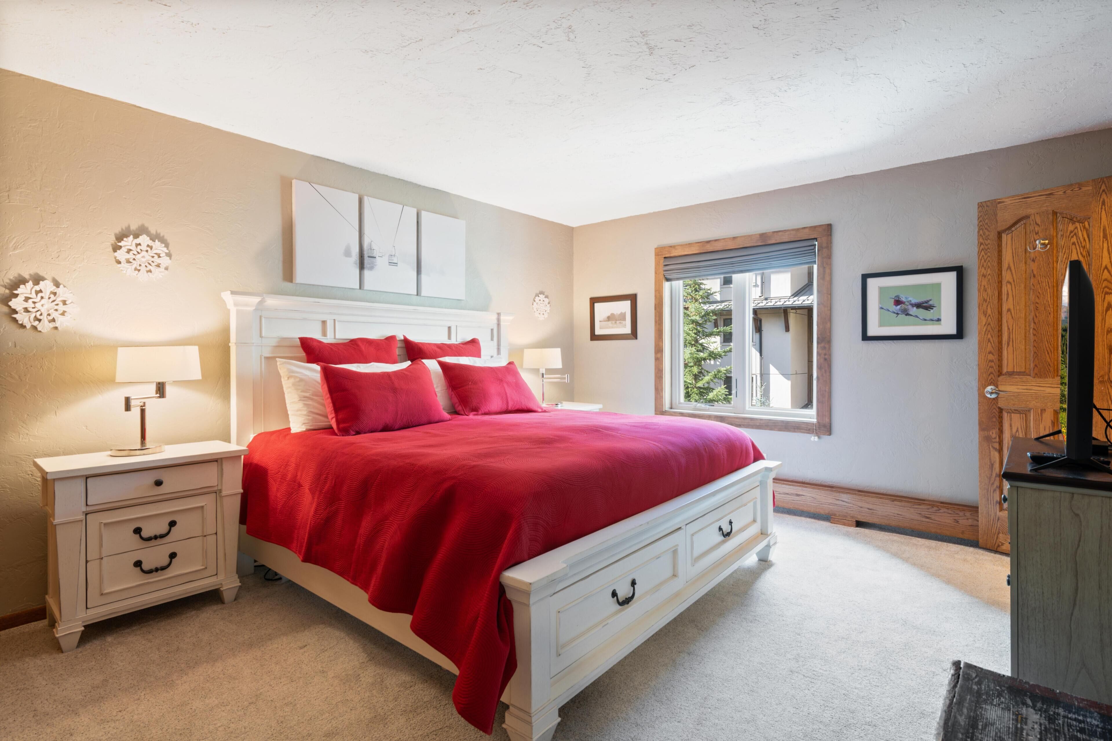 A bedroom with a white bed, red bedding, a nightstand with a lamp, a wooden door, and a window with a view.