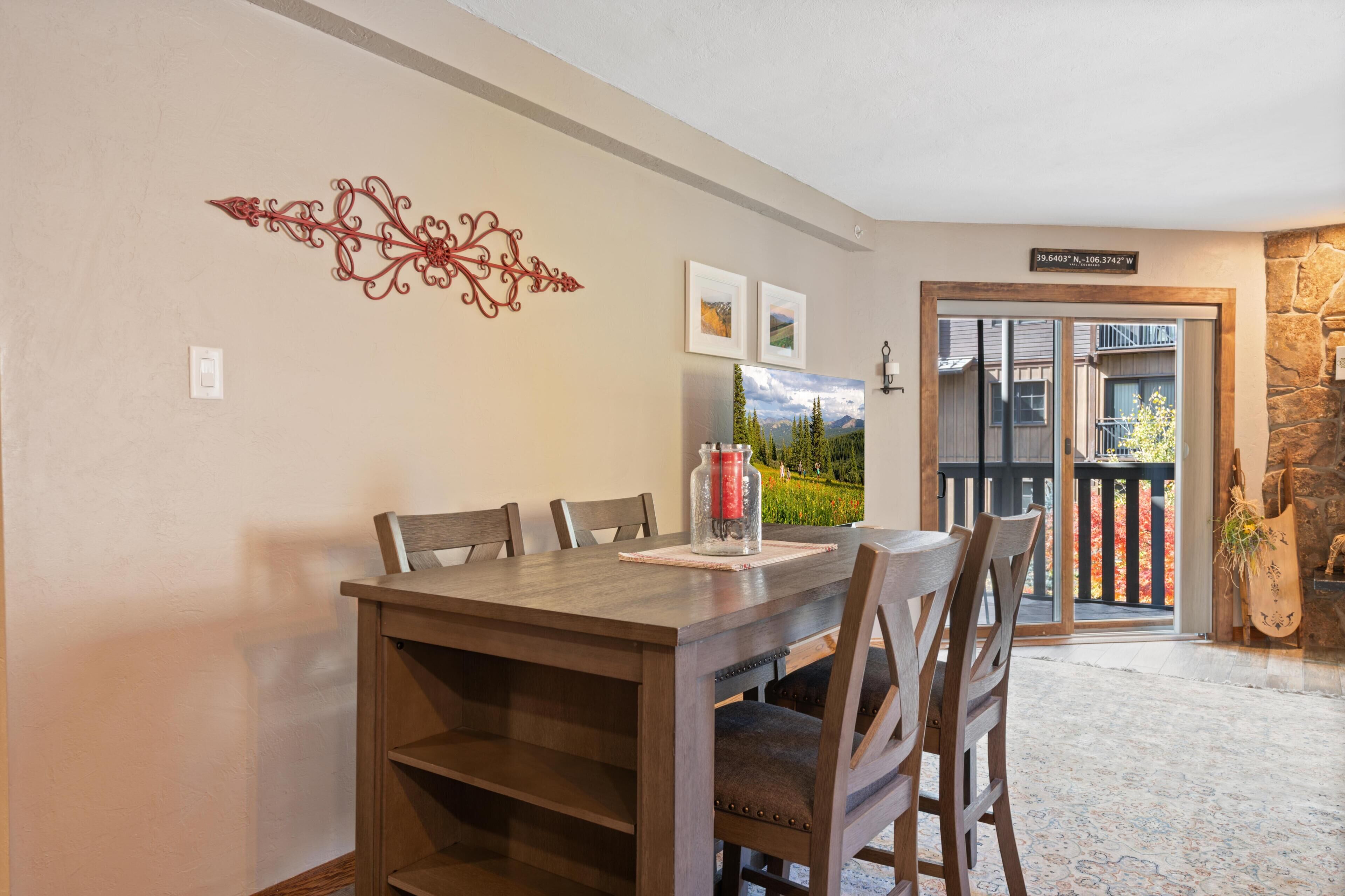 A dining area with a wooden table and chairs, a decorative wall piece, framed pictures, and a view of the outside through a glass door.