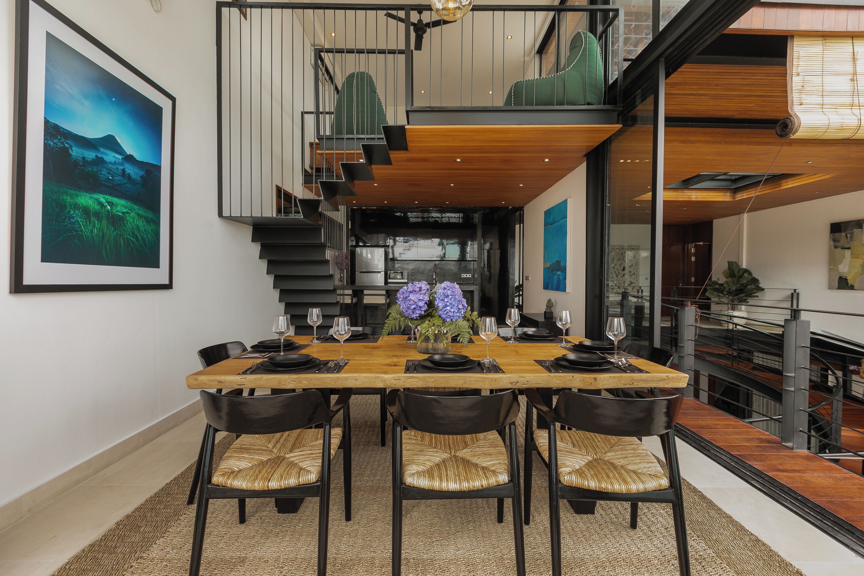 A dining area with a wooden table, black chairs, and a view of the kitchen and upper floor.