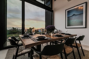 Comedor con mesa de madera dispuesta para seis comensales, ventanas amplias y un cuadro enmarco con un paisaje.