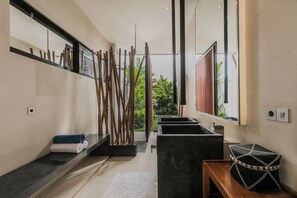 Un baño moderno con un espejo grande, un separador de listones de madera, un lavamanos negro y un banco con un bolso tejido.