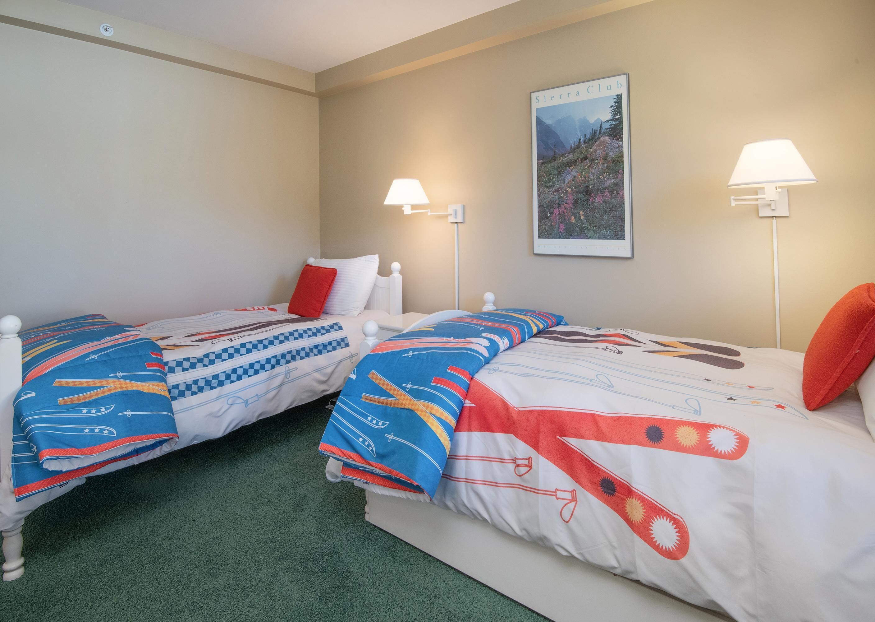 A hotel room with two single beds, each with a pillow and a blanket. There are two wall-mounted lamps and a framed picture on the wall.
