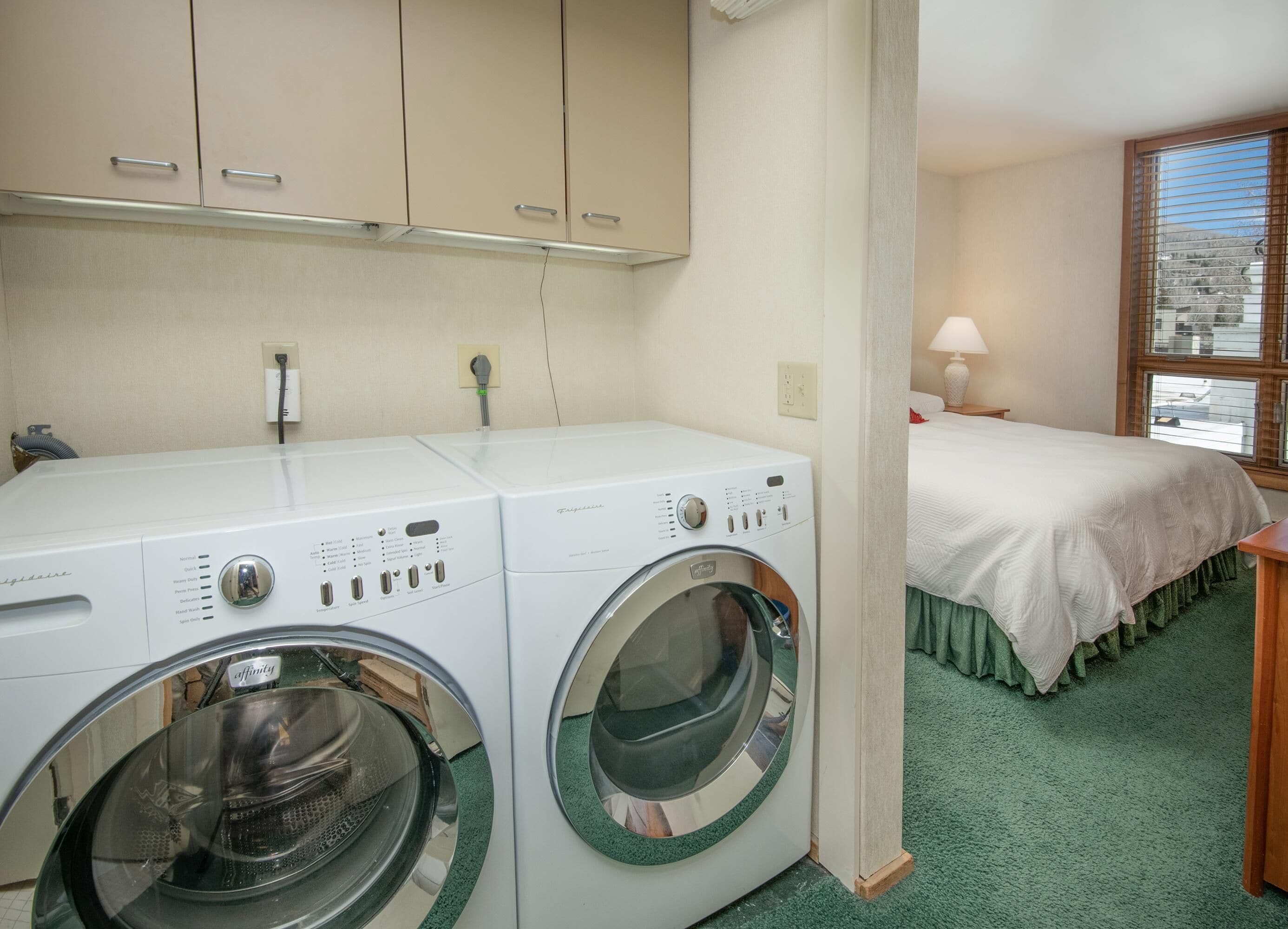 A laundry area with two front-loading washing machines, a bed with a green skirt, and a window with blinds.