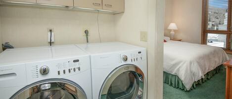 A laundry area with two front-loading washing machines, a bed with a green skirt, and a window with blinds.