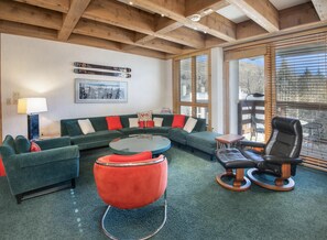 A cozy living room with a sectional sofa, a red ottoman, a leather chair, a glass coffee table, and wooden beams on the ceiling. - Treetops 6A, 3BD Ski-in Ski-out Condo (Vail)