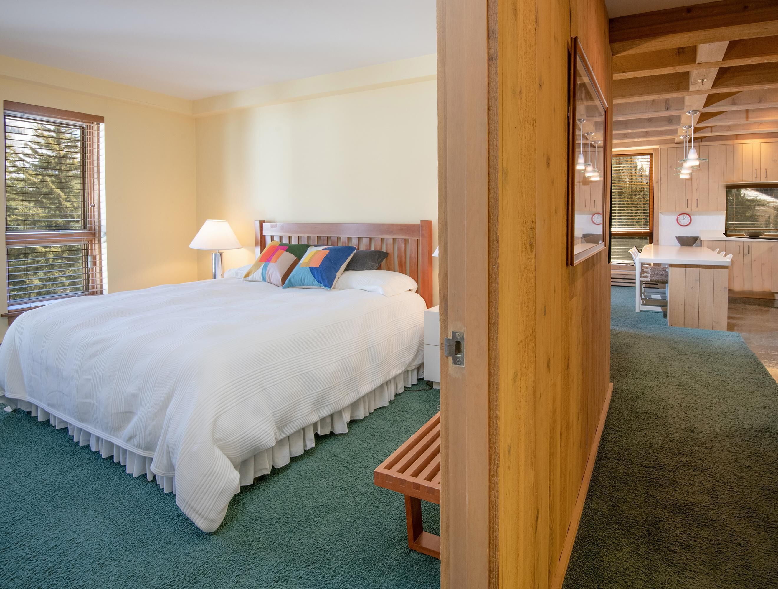 A bedroom with a large bed, a wooden headboard, a bedside table, a lamp, and a view of the kitchen area.