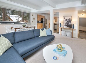 A modern living room with a blue sofa, a white coffee table, and a view of a kitchen and dining area. - Gore Circle Chalet, 4BD 3BA Private East Vail Home (Vail)