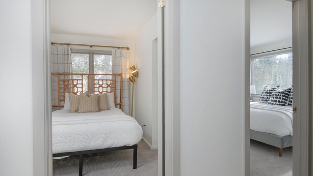 Un dormitorio con una cama, una ventana con cortinas, una lámpara de noche y vista a otra habitación.