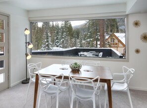 A dining area with a table, chairs, a lamp, and a view of a snowy landscape through a large window. - Gore Circle Chalet, 4BD Private East Vail Home (Vail)
