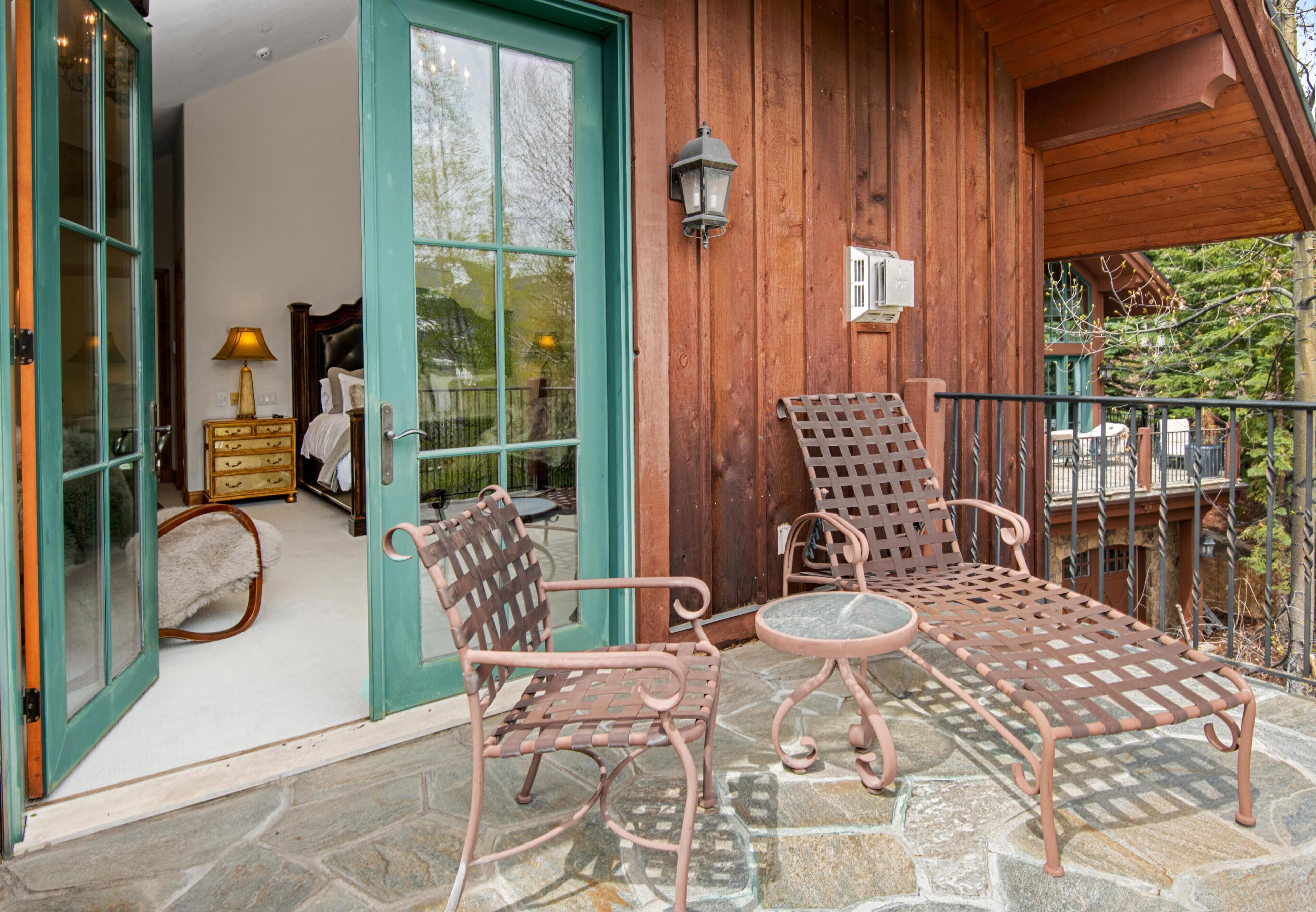 A wooden exterior with a stone patio, two wicker chairs, a metal table, and a glass door leading inside.