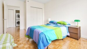 Makuuhuoneessa on sänky, yöpöytä, lamppu, pesukone ja ovi, joka johtaa toiseen huoneeseen.