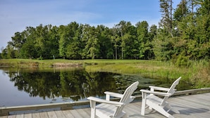 A wooden deck with two Adirondack chairs facing a calm pond surrounded by lush greenery. - Bayside Resort Escape: 5BR Coastal Home -Golf, Pools, Restaurants & Beach Access (Selbyville)