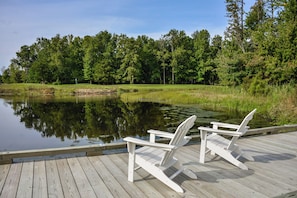A wooden deck with two Adirondack chairs facing a calm pond surrounded by lush greenery.