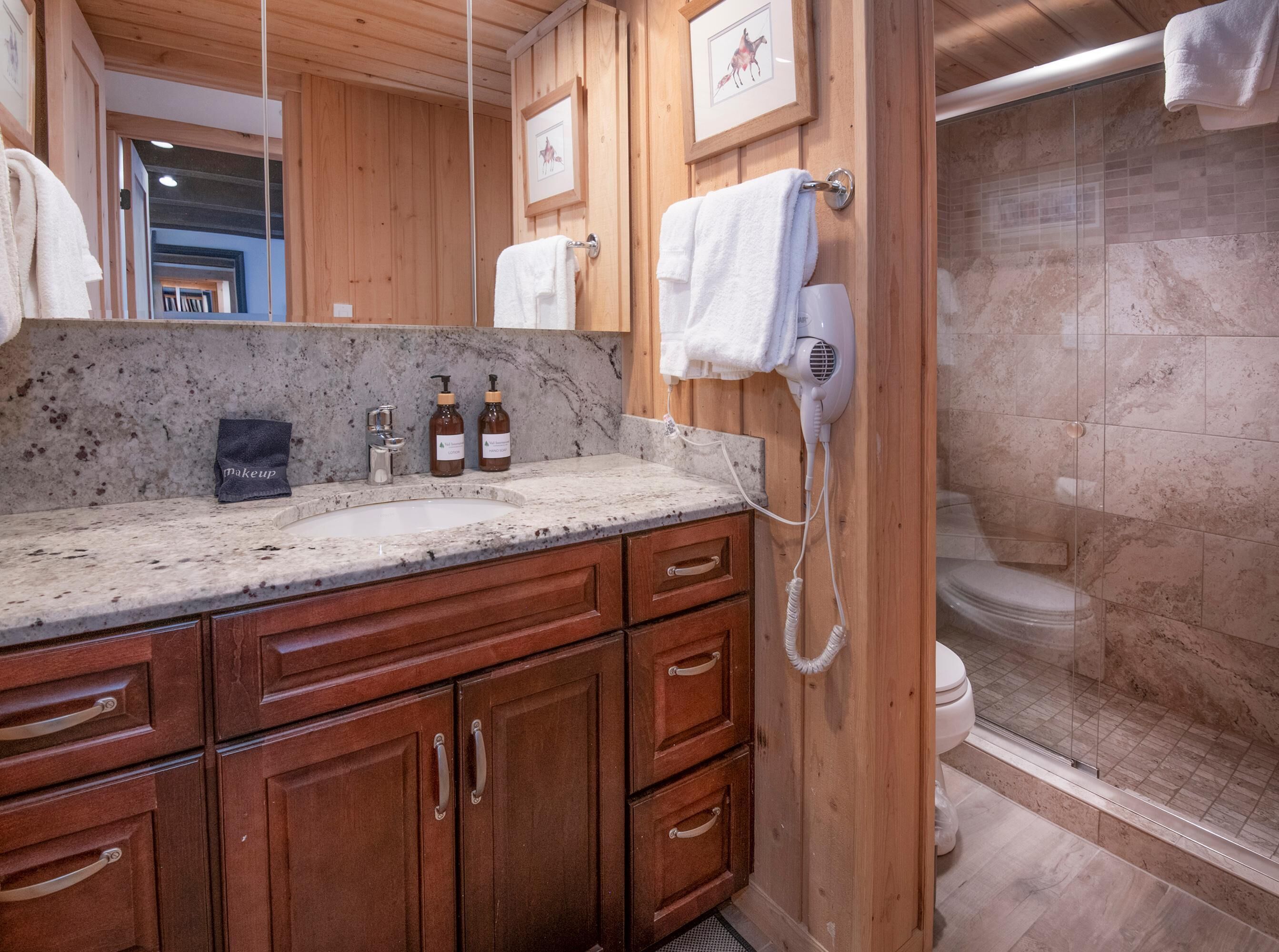 A bathroom with wooden cabinets, a marble sink, a hairdryer, and a shower with a glass door.