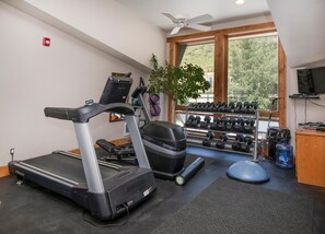 Ein Fitnessraum mit Laufband, Kurzhanteln, Wasserspender und Deckenventilator.