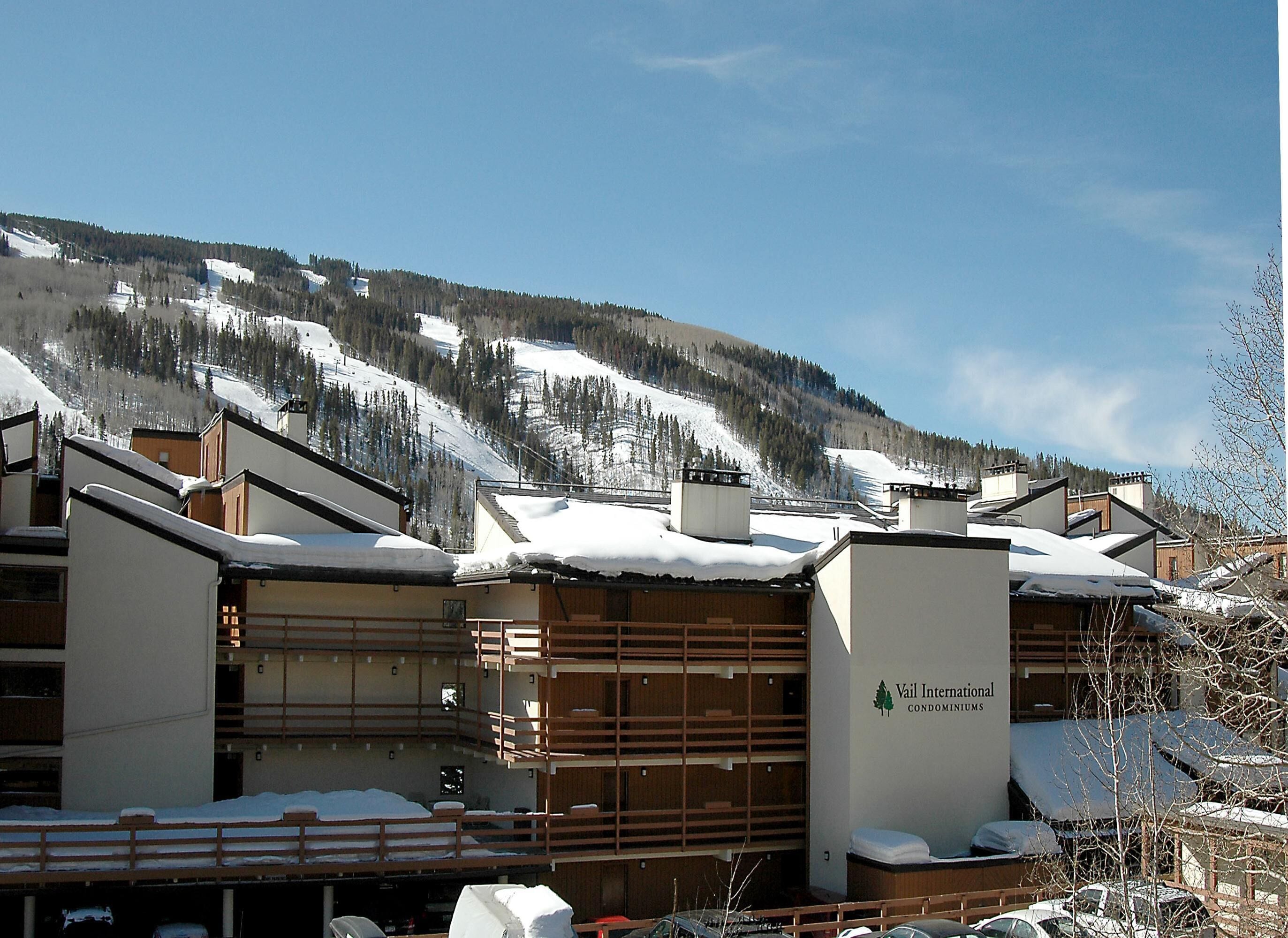 Un edificio de varios pisos con un letrero que indica Vail International Condominiums, rodeado de montañas nevadas y un cielo despejado.