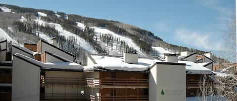A multi-story building with a sign for Vail International Condominiums, surrounded by snow-covered mountains and a clear sky.