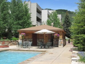 A pool area with a covered seating area, stone walls, and a view of a multi-story building in the background. - Sun Vail 13e, 2bd, 2BA Condo With Vail Mountain Views (Vail)