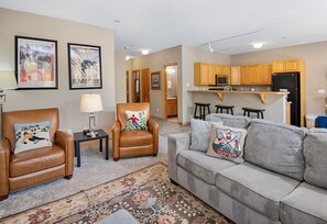 A living room with a sofa, armchair, and coffee table, a kitchen with wooden cabinets and appliances, and a hallway leading to another room. - Sun Vail 13e, 2bd, 2BA Condo With Vail Mountain Views (Vail)