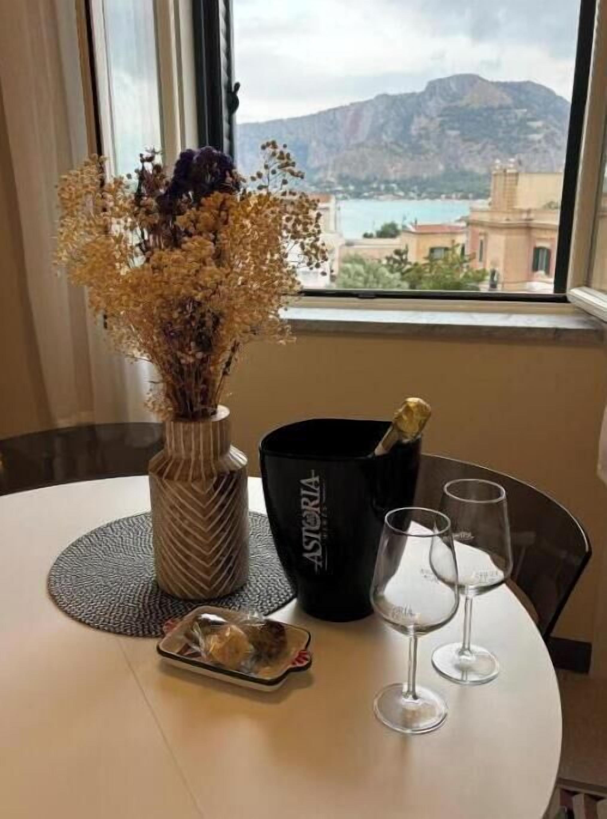 A table with a vase of dried flowers, a bottle, two glasses, and a small plate with a pastry.