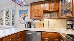 A kitchen with wooden cabinets, stainless steel appliances, a window with a view of trees, and a poster on the wall. - Westwind 209 1bd, 1BA Platinum Condo in Lionshead Village (Vail)