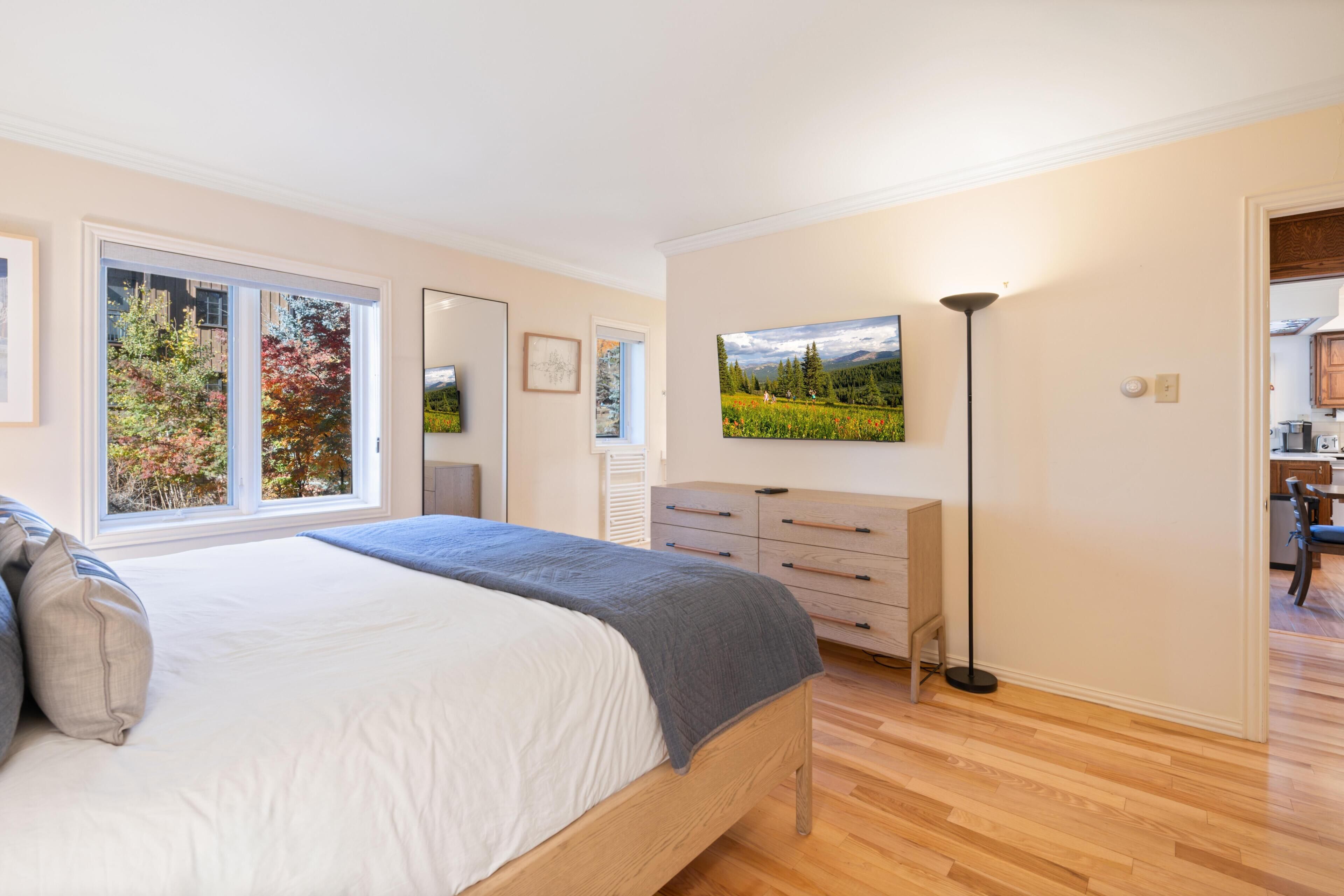 Een slaapkamer met een bed, een kaptafel, een nachtkastje, een lamp en een raam met uitzicht op bomen.