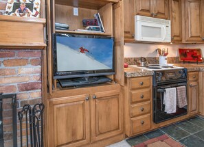 A kitchen with wooden cabinets, a microwave, a stove, a television, and a brick wall. - Wall Street 305, 1BD + Loft Condo in Vail Village (Vail)