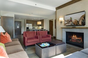 A modern living room with a fireplace, a television, and a kitchen in the background. - Riverfront Resort & Spa 421, 2BD Condo With Mtn Views (Avon)