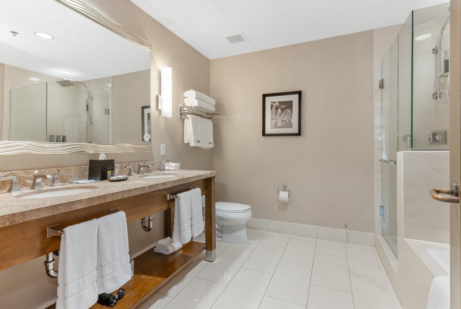 A modern bathroom with a large mirror, dual sinks, a toilet, a glass shower enclosure, and a framed picture on the wall.