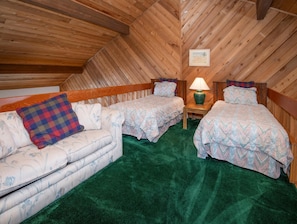Ein Zimmer mit geneigter Decke, zwei Einzelbetten mit passenden Kopfteilen, ein Sofa mit kariertem Kissen, ein kleiner Tisch mit Lampe und ein gerahmtes Bild an der Wand.