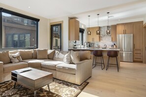 A modern living room with a sectional sofa, a coffee table, and a kitchen area with a refrigerator, bar stools, and pendant lights. - One Riverfront 106, 2BD Townhome With Beaver Creek Views (Avon)