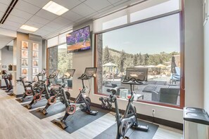 A gym with stationary bikes, a TV screen, and a view of a pool area. - One Riverfront 106, 2BD Townhome With Beaver Creek Views (Avon)