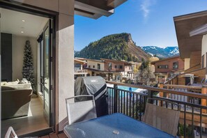 A balcony with a table, chairs, a grill, and a view of a mountain and buildings. - One Riverfront 106, 2BD Townhome With Beaver Creek Views (Avon)