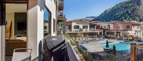A balcony with a grill, a pool area with lounge chairs, and a mountain backdrop.