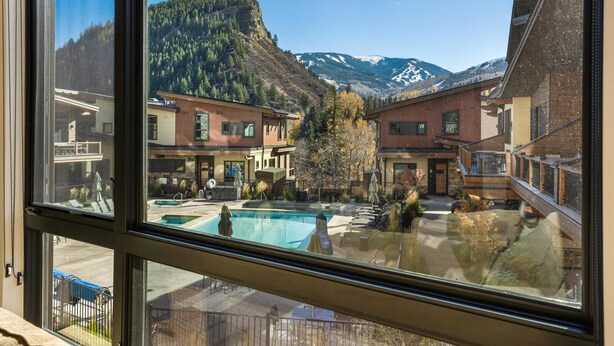 Blick durch ein Fenster auf einen Hotelinnenhof mit Pool, Außenbereich zum Sitzen und einem Bergpanorama.