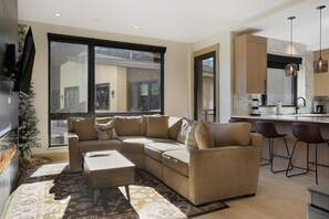 A modern living room with a sectional sofa, a coffee table, and a large window providing a view of the city. - One Riverfront 106, 2BD Townhome With Beaver Creek Views (Avon)
