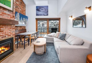 A cozy living room with a fireplace, a sofa, a dining table, and a view through a glass door. - Wall Street 307, 1BD + Loft Condo in Vail Village (Vail)