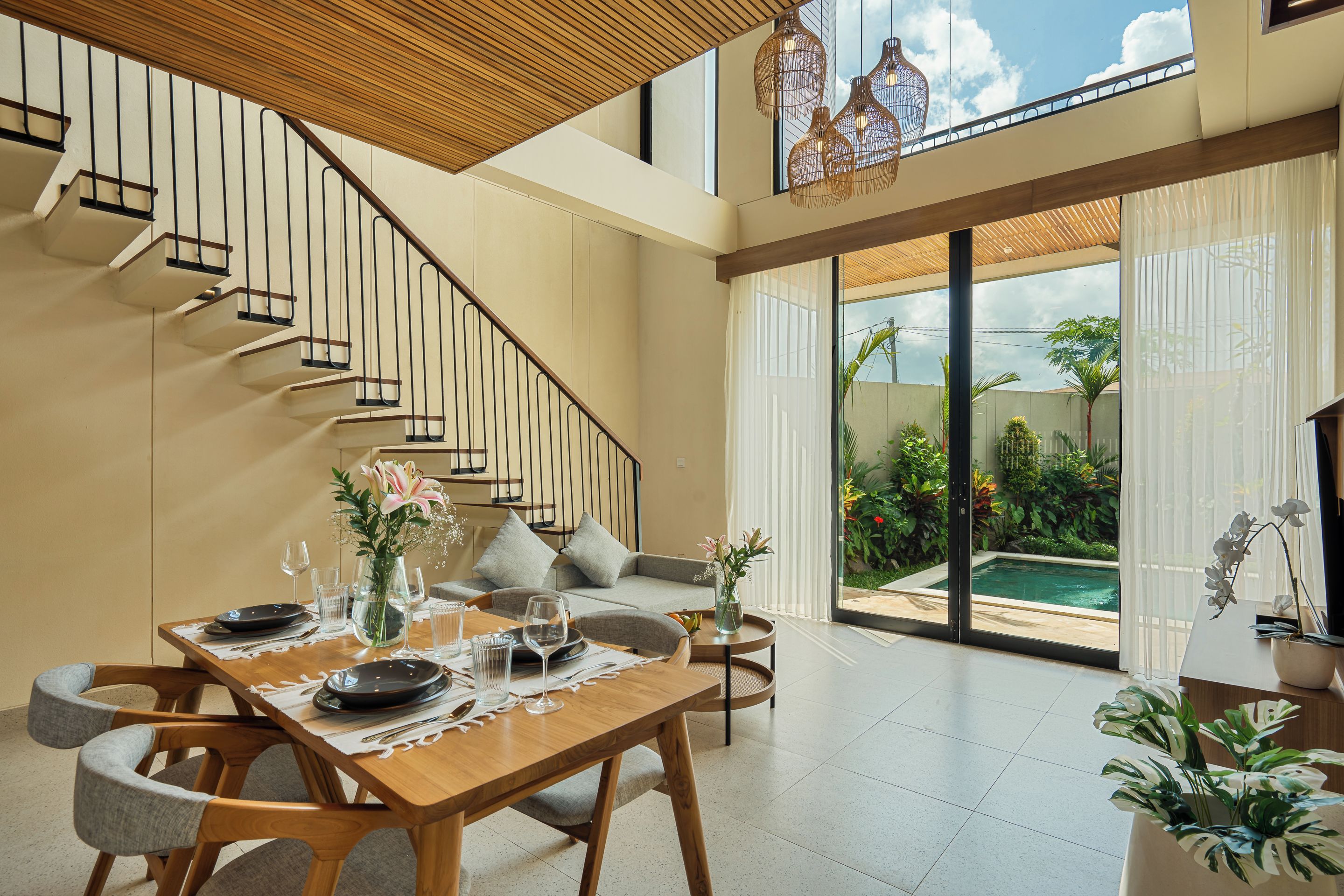 A modern interior with a wooden staircase, a dining area with a table set for two, and a view of a swimming pool through large glass doors.