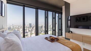 Ein modernes Hotelzimmer mit einem großen Bett, einem Holztischchen mit einer Serviertafel, einem an der Wand montierten Fernseher und einem Blick auf die Stadt durch große Fenster.