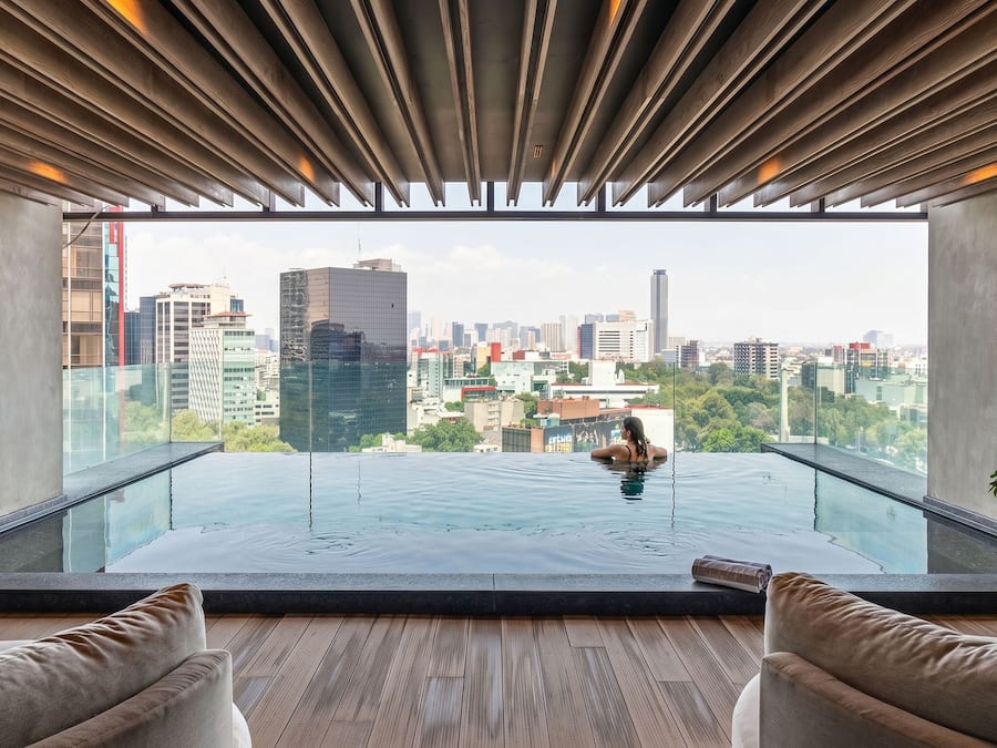Piscina infinita moderna no terraço com vista para a cidade, piso de madeira e dois sofás.