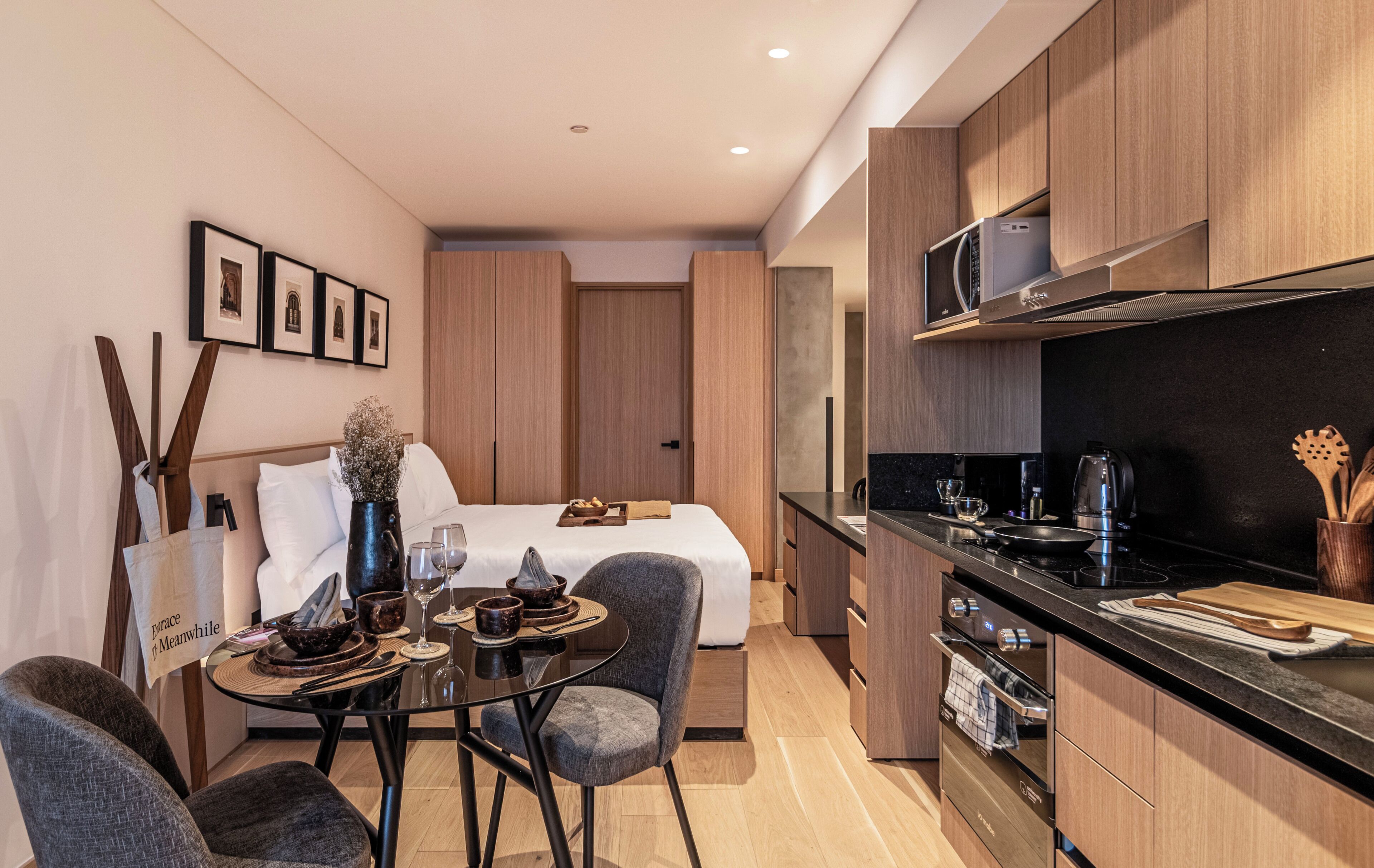 Una habitación de hotel moderna con una cama, una mesa de comedor con sillas, una cocina con microondas y una pequeña mesa con un jarrón y un bolso.