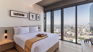 Une chambre d’hôtel moderne avec un grand lit, une table de chevet en bois, un plateau de petit-déjeuner et une vue sur la ville à travers de grandes fenêtres.