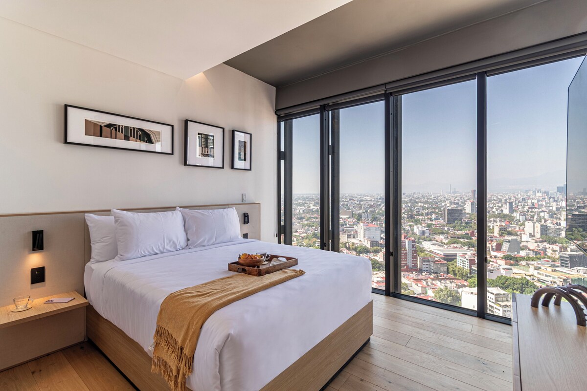Camera d'albergo moderna con un letto grande, un comodino in legno, un vassoio con prodotti per la colazione e vista sulla città attraverso ampie finestre.