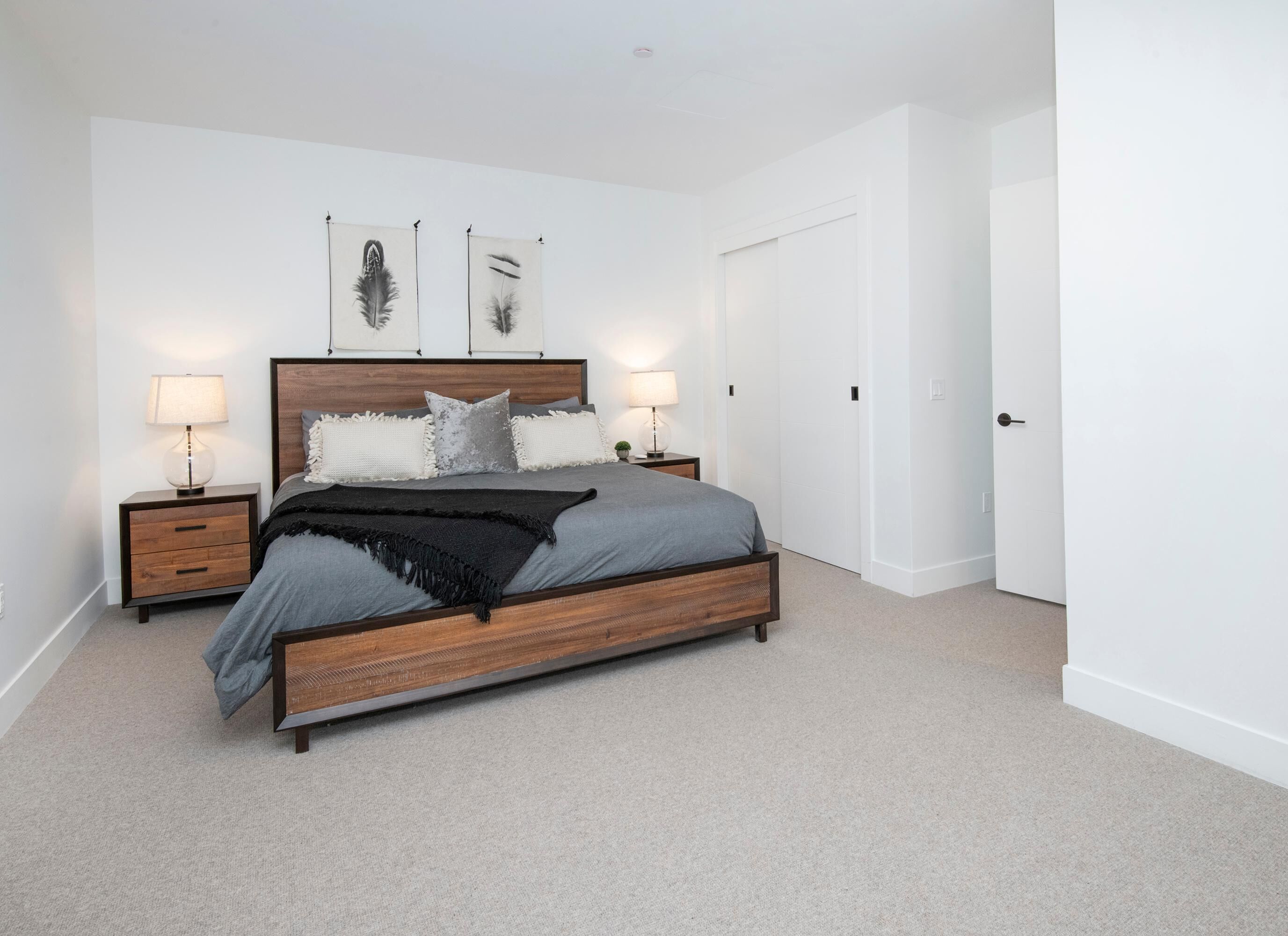 A bedroom with a wooden bed, two bedside tables with lamps, a grey blanket, and two feather wall art pieces.