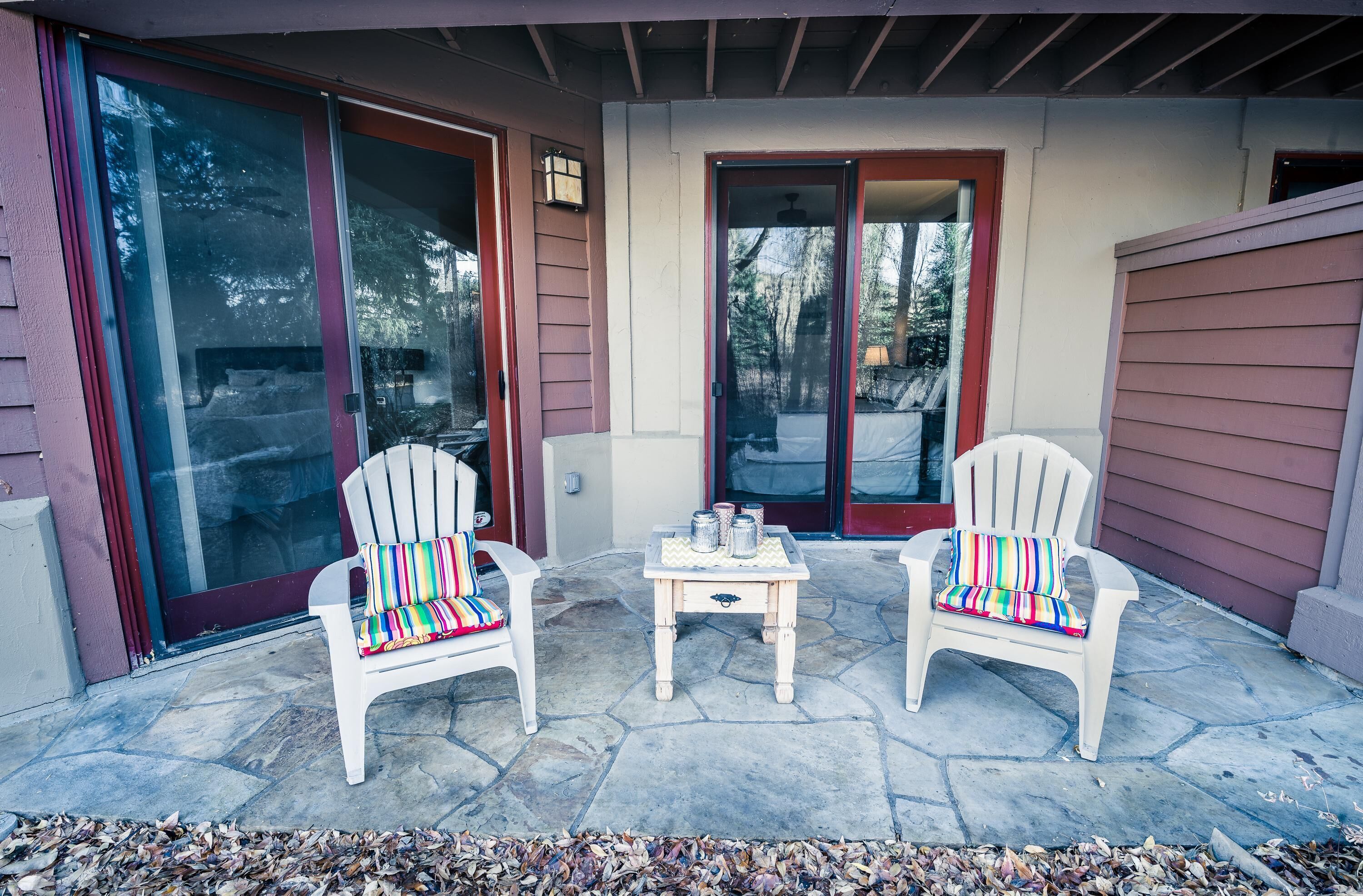 A covered patio with two Adirondack chairs, a small table with a decorative top, and a sliding glass door.