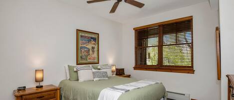 A bedroom with a bed, a nightstand, a wall-mounted lamp, a ceiling fan, a window with blinds, and a poster on the wall.