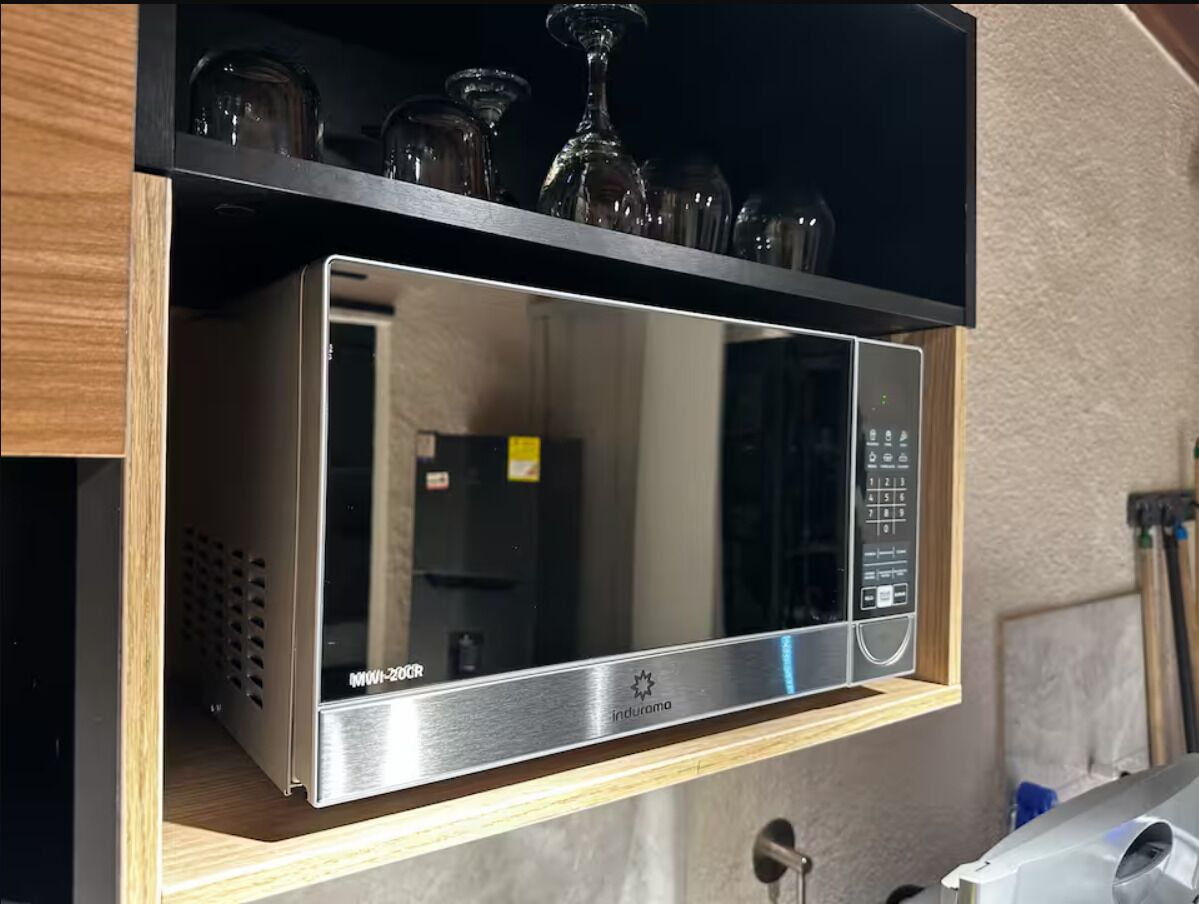 Forno de microondas em aço inoxidável com painel de controle digital, posicionado sobre uma prateleira de madeira acima de uma bancada de cozinha.