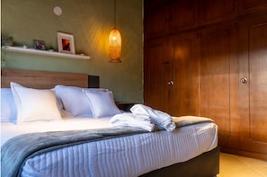 A neatly made bed with pillows and a folded towel, a wooden wardrobe, and a wall-mounted shelf with decorative items. - Modern Suite with Private Balcony • A/C • WiFi (Medellín)